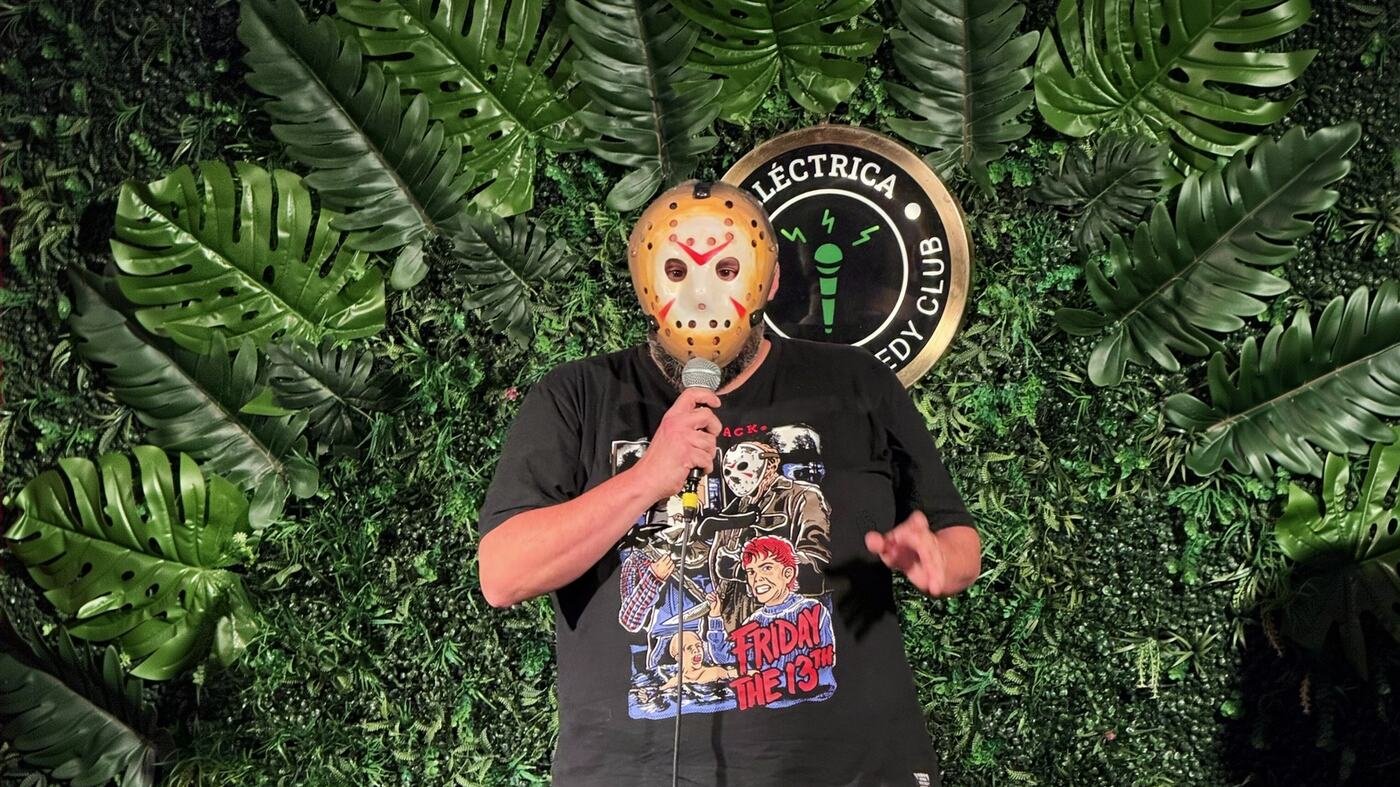 Maxi en el escenario de un club de stand up, con una máscara de hockey y el micrófono en la mano, frente a una pared de plantas