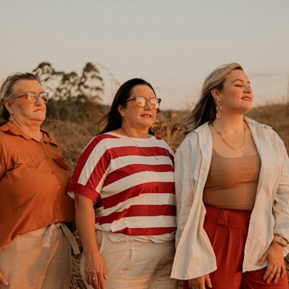 Tres mujeres adultas de pie en un campo al atardecer, mirando en distintas direcciones