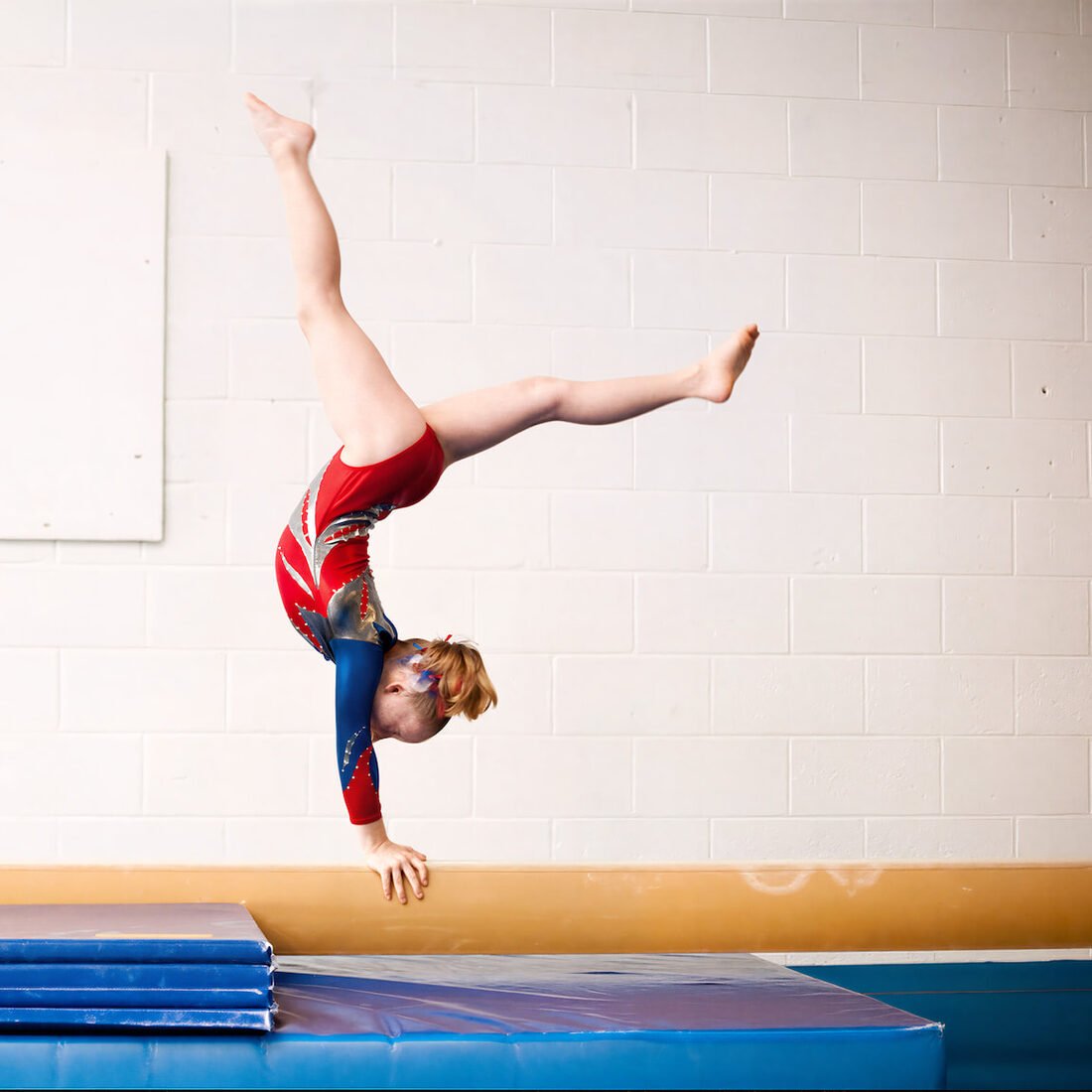 Una niña haciendo una vertical sobre una viga de gimnasia, con colchonetas debajo