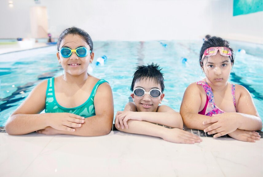 Tres niños con antiparras apoyados en el borde de una pileta durante una clase de natación