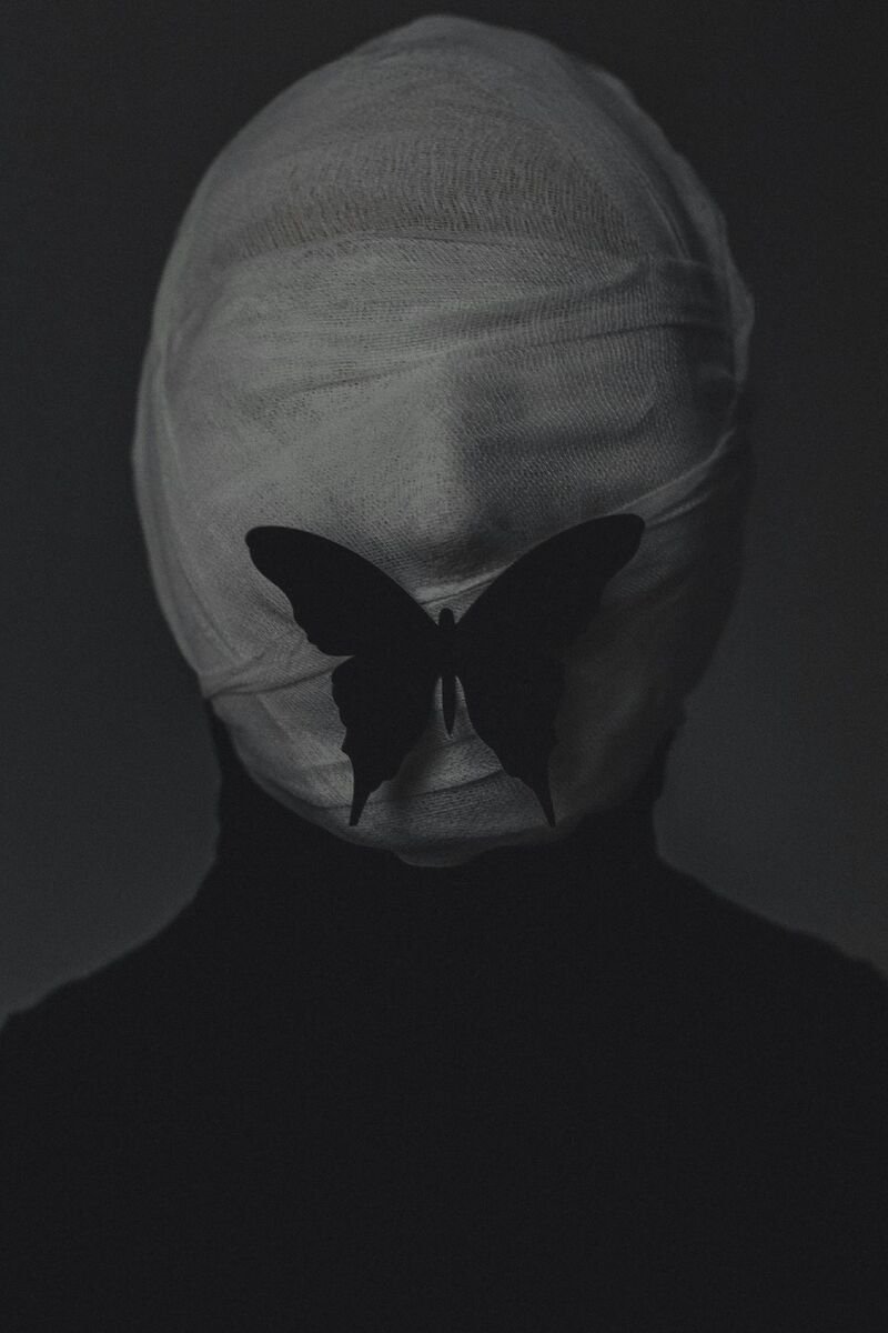 Retrato en blanco y negro de una persona con la cabeza vendada y una mariposa negra sobre la cara